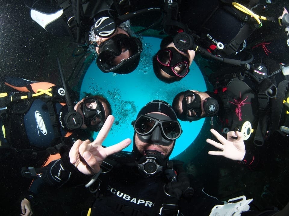 חמישה צוללים בתוך ספינת טילים צפים בצורת עיגול ומסתכלים למטה אל העדשה